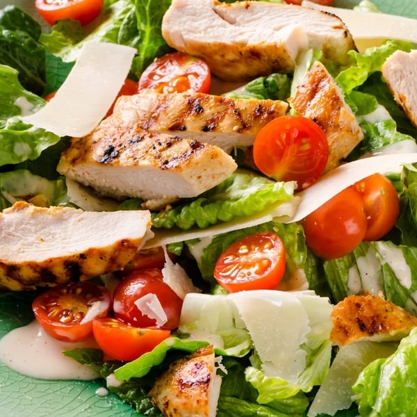 Grilled chicken Caesar salad with romaine lettuce, cherry tomatoes, parmesan, and dressing on a green plate.