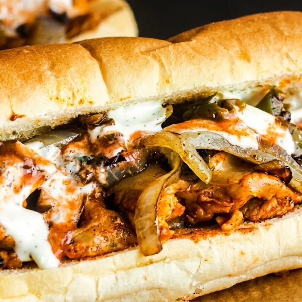 Close-up of a chicken cheesesteak sandwich with melted cheese, peppers, onions, and sauce on a bun.
