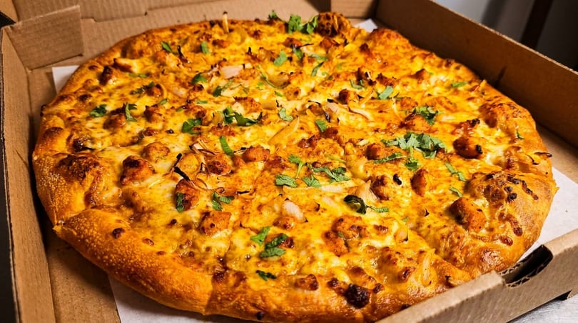 A whole chicken tikka pizza with cilantro and onions in a cardboard delivery box.